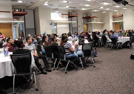 Attendees sit at round tables in a large event space during a special ceremony hosted by Rio Salado College for its High School ACE and Adult ACE programs. The room is filled with diverse students, families, and supporters attentively watching the stage (not visible in the image). The tables are decorated with white cloths and centerpiece arrangements. A projected display on the wall references the year “2025,” suggesting a recognition or graduation event.