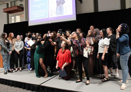 A lively group of students and staff gather on stage at a Rio Salado College event, smiling and posing for a group selfie. Several people in front hold up smartphones to capture the moment, while others cheerfully join in. One woman in a sparkling green gown leads the front row, with certificates visible in the hands of a few participants. Behind them, a large screen displays a student testimonial and image. The joyful, informal atmosphere contrasts with the formal stage setting, celebrating a milestone.