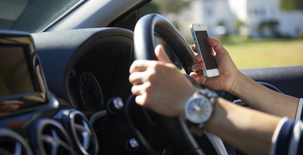 Image of a car steering wheel with a driver's one hand on wheel and other hand on a cell phone