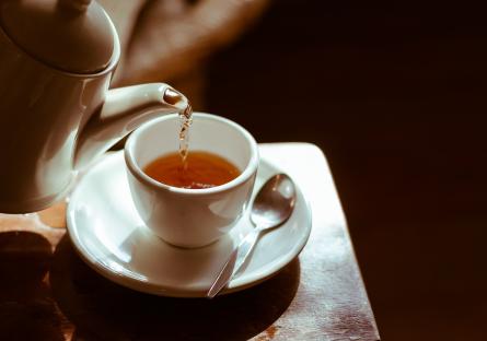 A teapot pouring tea into a cup on a saucer with a spoon on a table