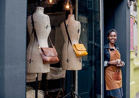 A female entrepreneur stands outside her designer women's shop, with a cup of coffee.