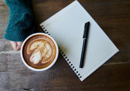 A hand holding a latte next to a notebook with a pen